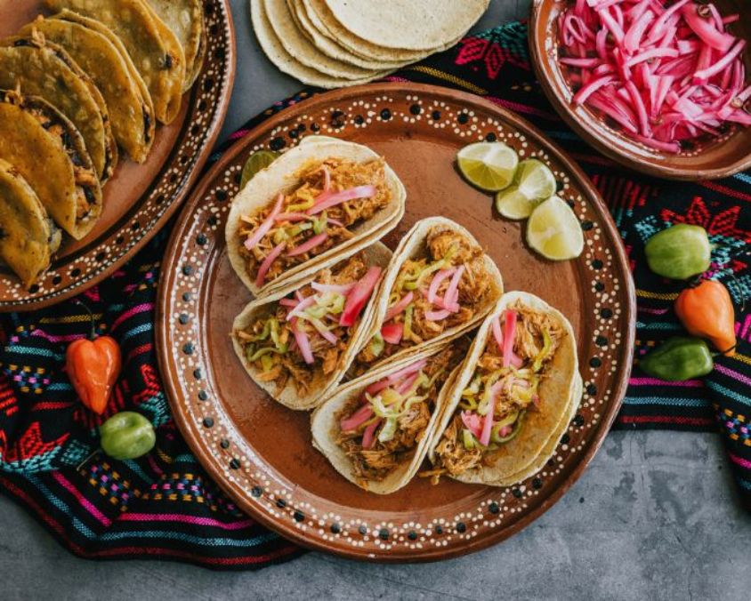 A colourful Mexican-style food spread featuring four soft tacos filled with shredded meat, sliced pickled onions and fresh toppings on a decorative clay plate. Lime wedges sit beside the tacos, with extra corn tortillas, a bowl of pickled onions and additional tacos arranged around the scene on a patterned textile.