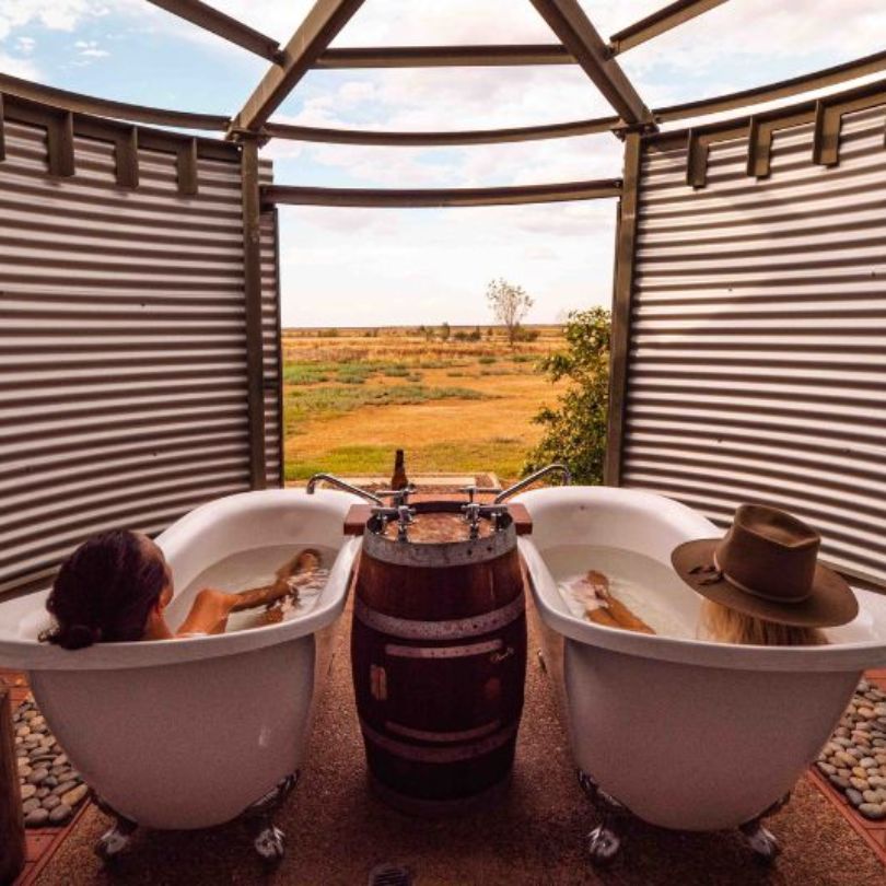 Two visitors soaking in outdoor clawfoot bathtubs at Julia Creek, with a scenic rural landscape in the background and a rustic setup featuring a shared wooden barrel faucet.