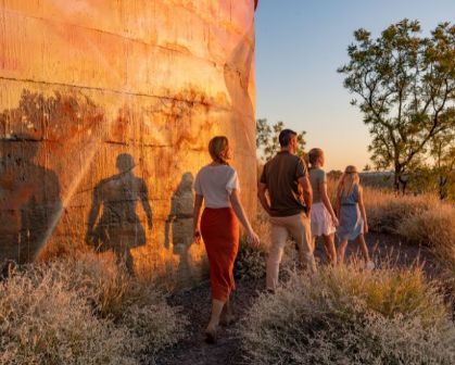 Picture of Visitors walking beside the Cloncurry Water Tank Mural at sunset, with long shadows cast on the cylindrical structure and outback scenery in the background.