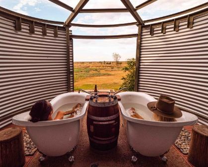 Picture of Two visitors soaking in outdoor clawfoot bathtubs at Julia Creek, with a scenic rural landscape in the background and a rustic setup featuring a shared wooden barrel faucet.