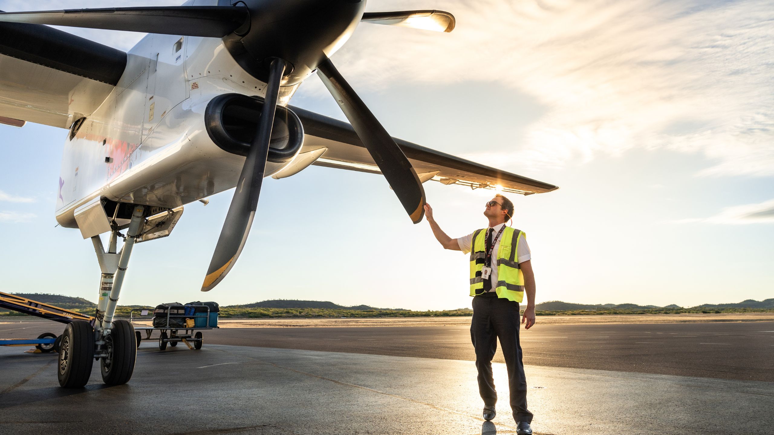 Person in high-visibility vest photographing and inspecting a propeller aircraft on the tarmac at Mount Isa Airport during sunrise or sunset.